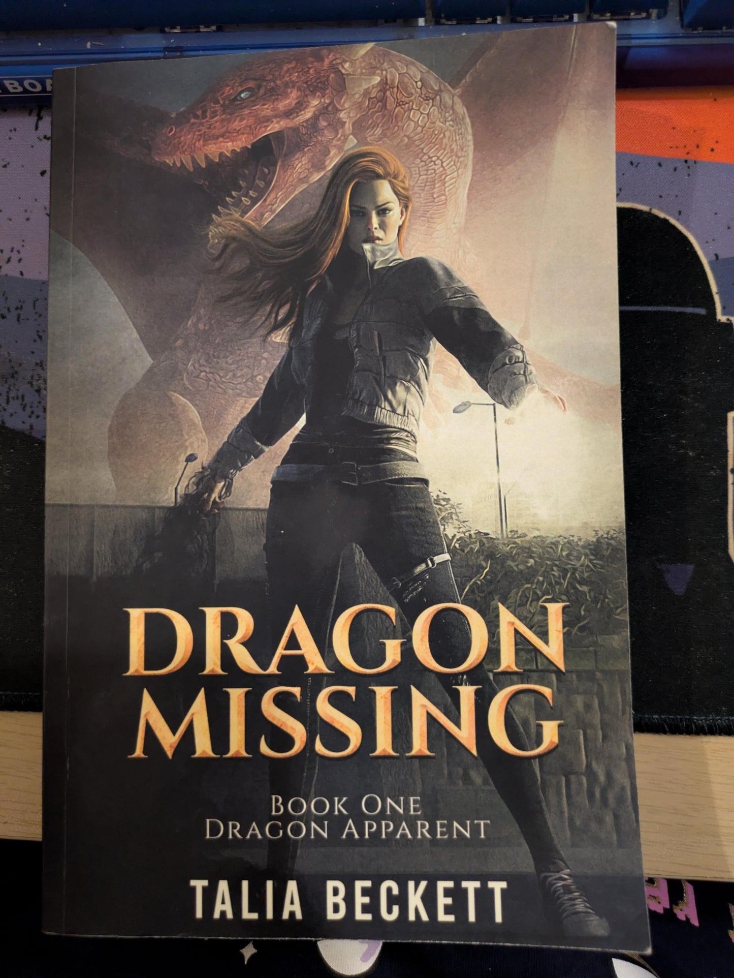 Book cover for 'Dragon Missing' by Talia Beckett, featuring an illustration of a woman in a dark outfit with a large dragon in the background, photographed against a dark surface.