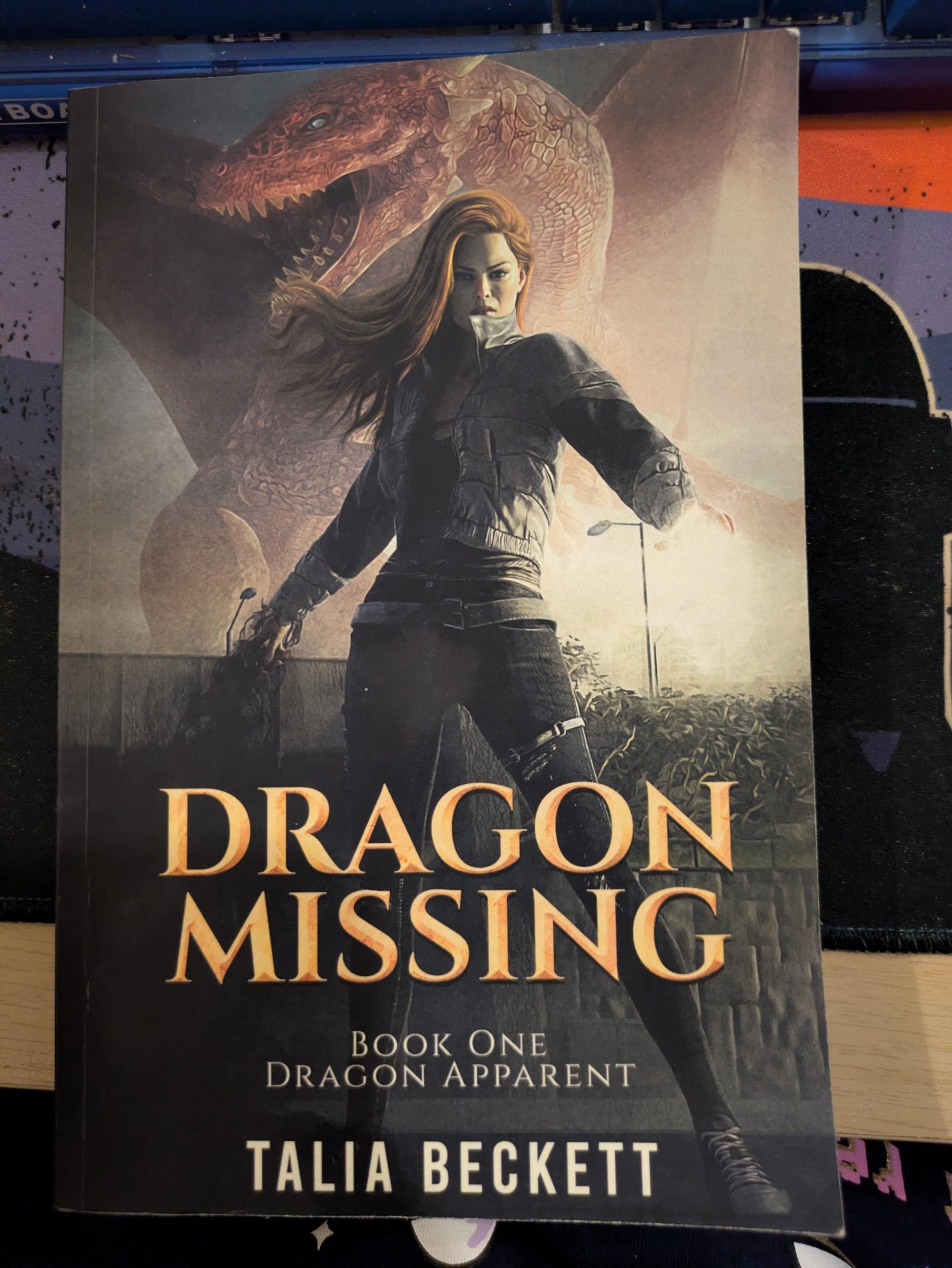 Book cover for 'Dragon Missing' by Talia Beckett, featuring an illustration of a woman in a dark outfit with a large dragon in the background, photographed against a dark surface.