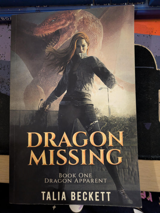 Book cover for 'Dragon Missing' by Talia Beckett, featuring an illustration of a woman in a dark outfit with a large dragon in the background, photographed against a dark surface.